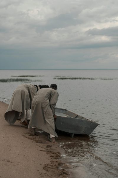 Two men in coats push a small boat onto a sandy beach under a cloudy sky, creating a serene seaside scene.